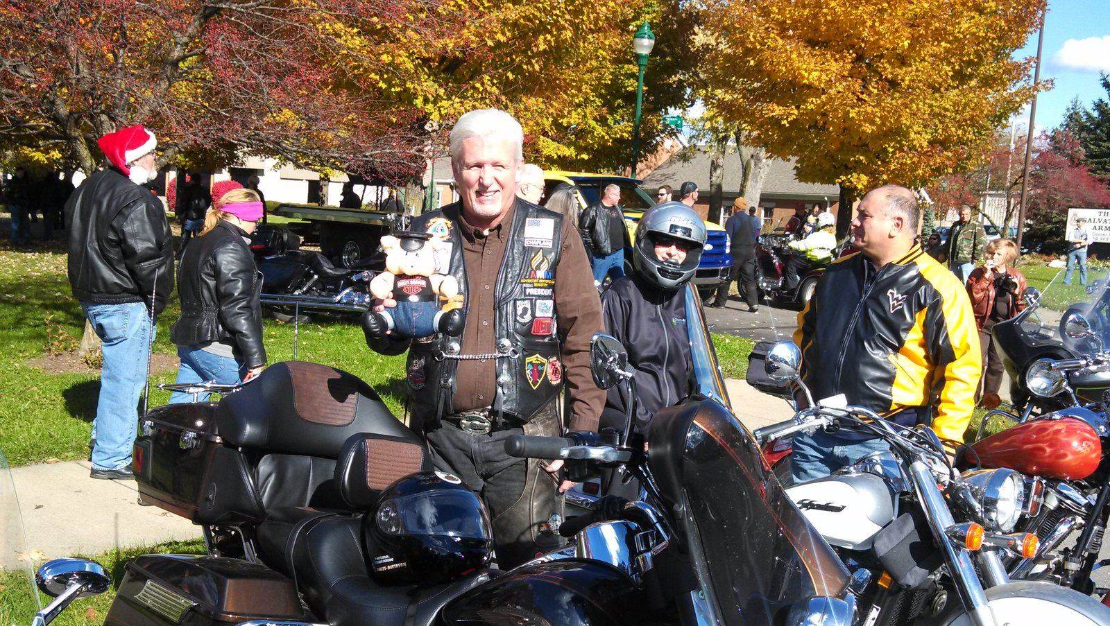 Tom Hughes holding a stuffed bear at a Toy Run, fall foliage and motorcycles behind him
