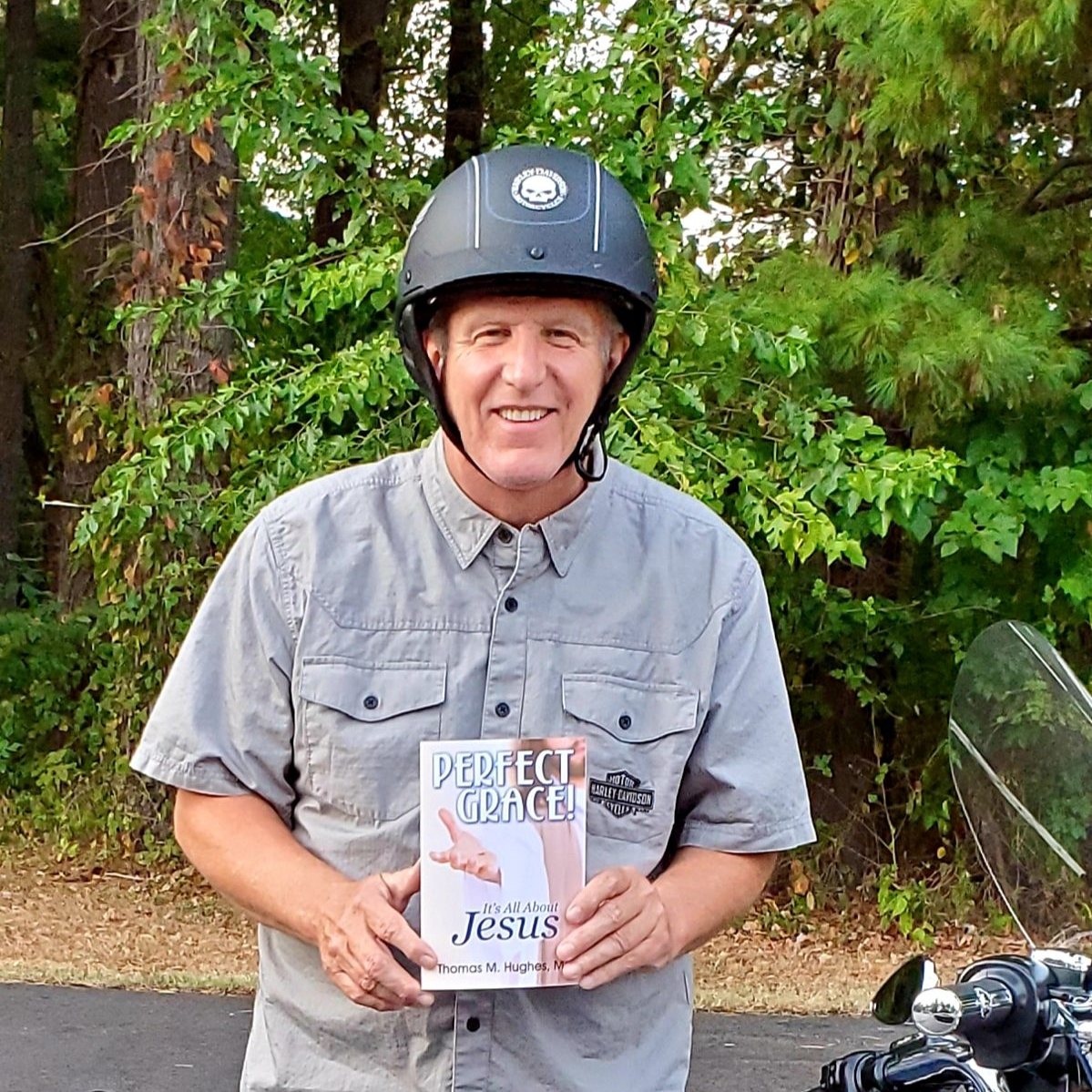 Tom Hughes in his helmet holding a copy of Perfect Grace! beside his Harley