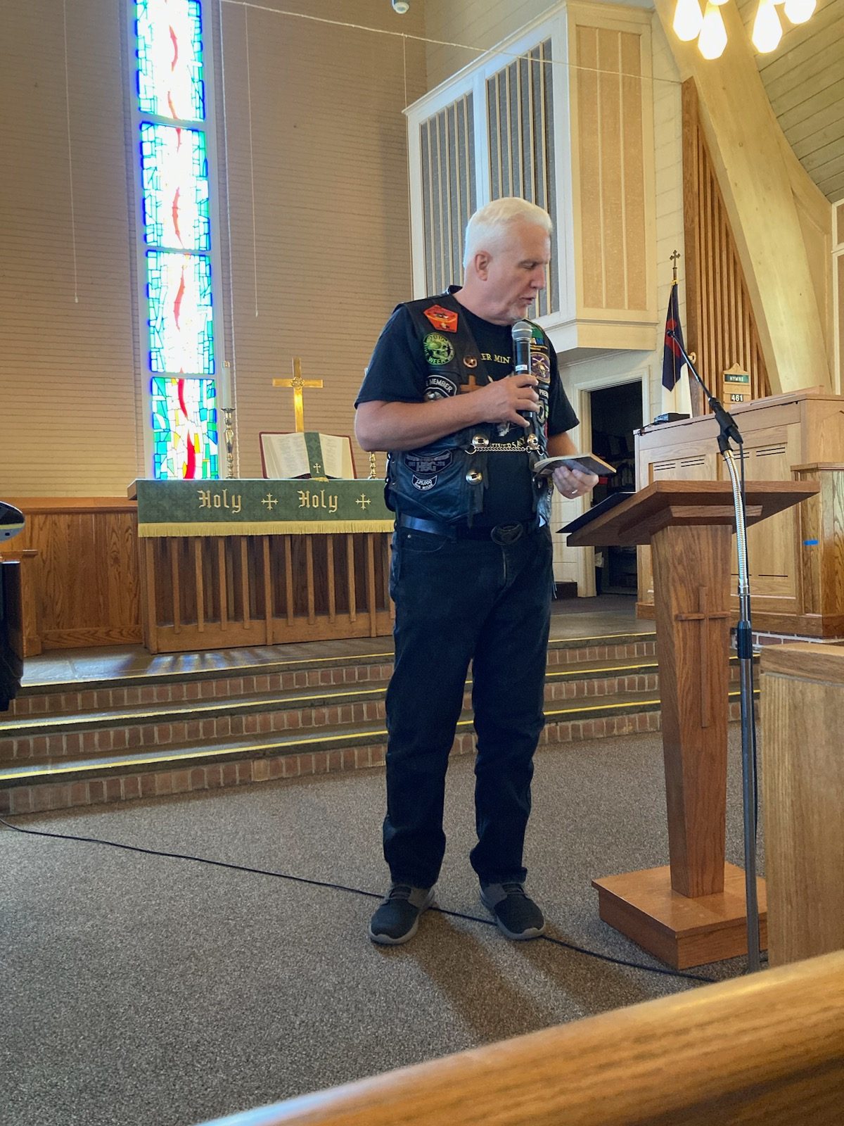 Pastor Tom Hughes preaching from the pulpit in his patched leather vest, stained glass behind him