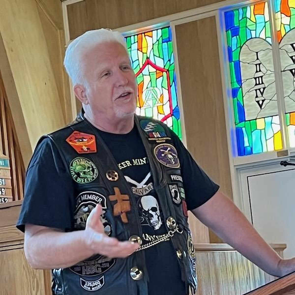 Pastor Tom Hughes in his patched leather vest, hands open, preaching in front of a stained-glass window