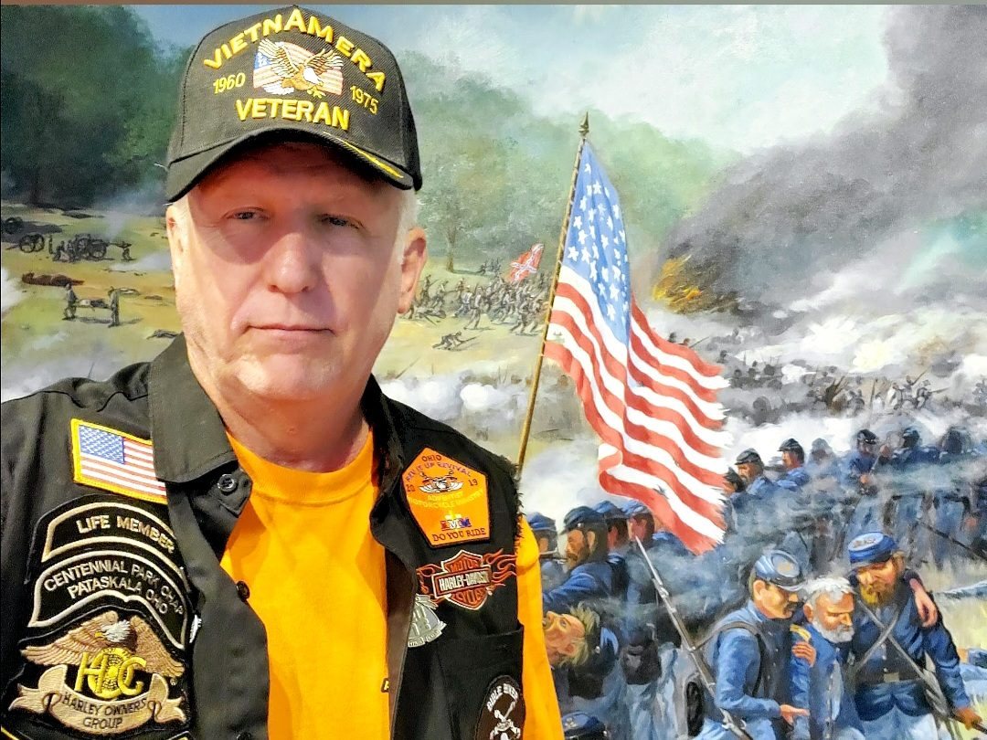 Tom Hughes in a Vietnam Era Veteran cap in front of a Civil War mural and the flag