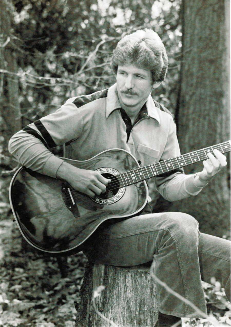 A young Tom Hughes playing acoustic guitar on a tree stump, black and white